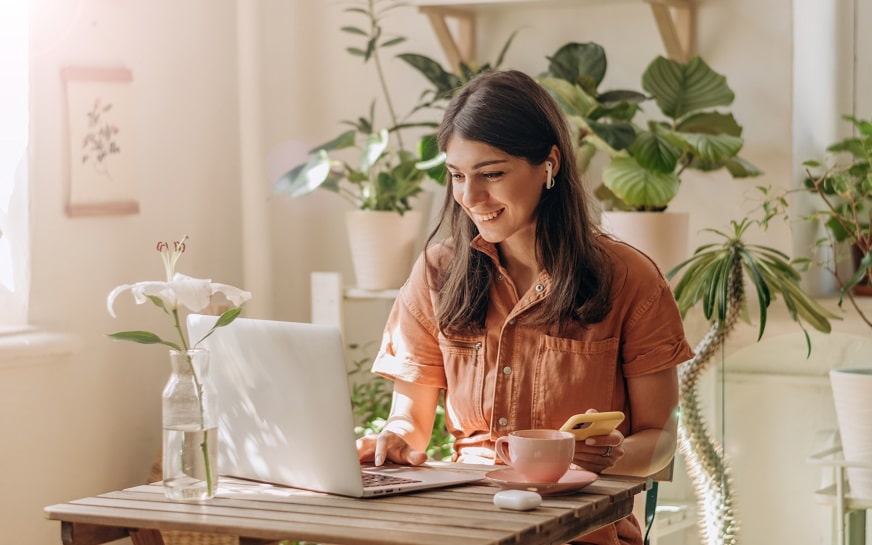 Positive young mixed race woman using a laptop and smartphone at home.Cozy home interior with indoor plants.Remote work, business,freelance,online shopping,e-learning,urban jungle concept